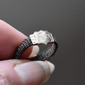 May include: A silver ring with a black braided band and a decorative engraved top. The top of the ring has a shield-shaped design with the initials "M.C." engraved on it.
