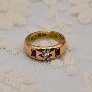 以下が含まれることがあります： 星形のデザインと中央にダイヤモンドが1つ付いたゴールドのリング。ダイヤモンドの両側に小さなルビーが2つセットされています。