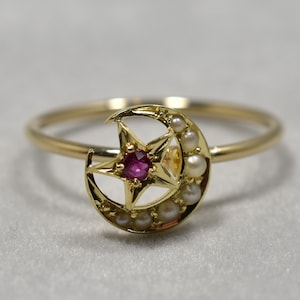 May include: A gold ring with a crescent moon design featuring a red ruby star in the centre and small white pearls around the moon.