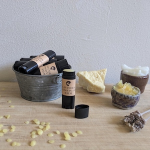 May include: A collection of natural skincare products, including lip balms, shea butter, and a scrub. The lip balms are in black tubes with labels that read "Lavender Lanolin Lip Balm." The items are arranged on a wooden surface.