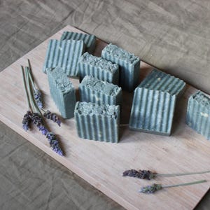 Puede incluir: Un conjunto de jabones rectangulares hechos a mano, de color verde apagado, dispuestos sobre una tabla de madera clara. Los jabones tienen una superficie texturizada y acanalada. Ramas de lavanda se colocan alrededor de los jabones.