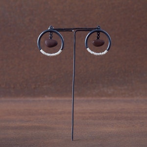 Könnte beinhalten: Ein Paar schwarze Creolen mit einem braunen Steinanhänger, der an jeder Creole hängt. Die Creolen sind mit einer Reihe weißer Perlen verziert.