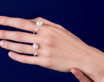 May include: Four silver rings, each featuring a white pearl or small clear stone, are shown on a hand against a dark blue backdrop. The rings have an open design, highlighting the gemstones.