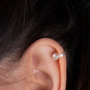 May include: A silver ear cuff featuring a small, round pearl. The cuff wraps around the ear's cartilage, creating a minimalist and elegant design. The pearl adds a touch of classic sophistication to the jewelry.