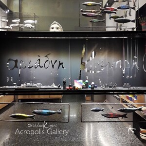 May include: A storefront window display with the text "Acropolis Gallery" and the Greek words "&alpha;&rho;&iota;ά&delta;&nu;&eta; &Kappa;ύ&pi;&rho;&iota;" below. The window is decorated with colorful fish sculptures hanging from the ceiling.
