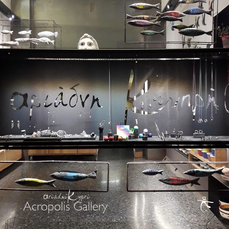 May include: A storefront window display with the text "Acropolis Gallery" and the Greek words "&alpha;&rho;&iota;ά&delta;&nu;&eta; &Kappa;ύ&pi;&rho;&iota;" below. The window is decorated with colorful fish sculptures hanging from the ceiling.