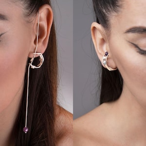 May include: Two silver earrings are displayed, one on each ear. The left earring features a geometric design with a long, dangling silver chain ending in a small, purple bead. The right earring is a silver hoop with a small, purple gemstone.