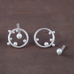 May include: Silver-toned earrings with a circular design, adorned with multiple small, white pearls. The earrings have a modern, minimalist aesthetic, with a single pearl dangling from the top of the circular design. The background is a solid, earthy brown.