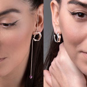 Puede incluir: Se muestran dos pendientes de plata. Uno es un pendiente largo y colgante con un diseño geométrico y una pequeña cuenta redonda morada en el extremo. El otro es un pendiente de aro pequeño y plateado con una forma abstracta.