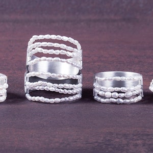 Op de afbeelding: Vier zilveren ringen met witte parelaccenten worden getoond tegen een donkerbruine achtergrond. De ringen hebben verschillende ontwerpen, met één met een brede band en meerdere parelstrengen, terwijl andere smallere banden en minder parelstrengen hebben.