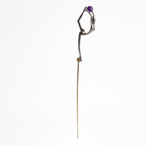 May include: A silver-toned sculpture of a person in a yoga pose, holding a purple gemstone. The sculpture is mounted on a thin gold rod.