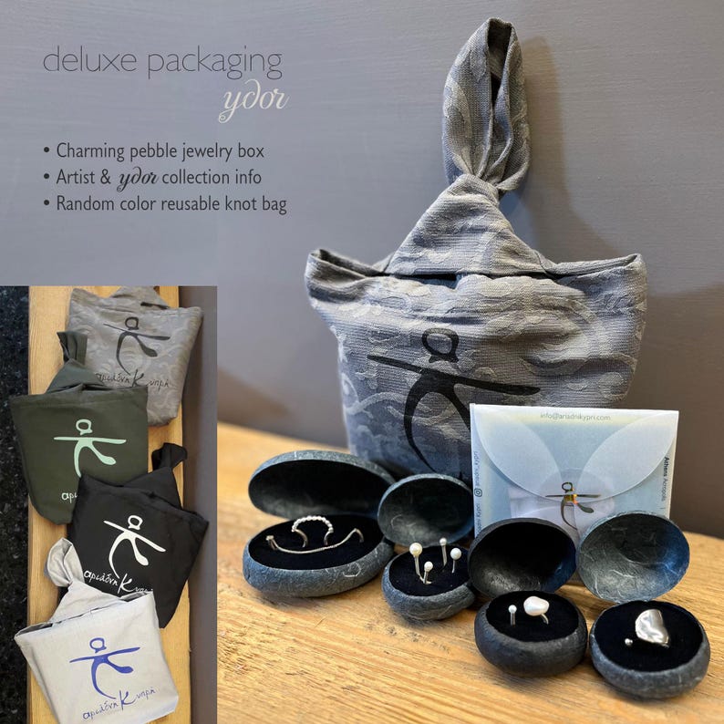 May include: A set of jewelry packaging featuring a gray reusable knot bag, a small white envelope, and several pebble jewelry boxes. The boxes are open and show various pieces of jewelry, including rings, earrings, and a pendant. The bag has the text "&alpha;&rho;&iota;ά&delta;&nu;&eta; &kappa;" printed on it.