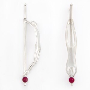 May include: A pair of silver earrings with a stylized human figure design and a red gemstone at the bottom.