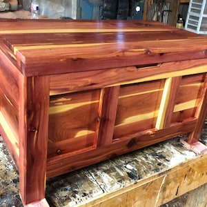 May include: A cedar wood chest with a hinged lid and a simple design. The chest is made of natural wood with a reddish-brown colour and has a light wood trim around the edges.