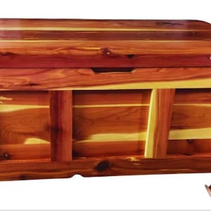 May include: A large wooden cedar chest with a hinged lid and a decorative front panel. The chest is made of natural cedar wood with a rich reddish-brown colour and a natural wood grain pattern.