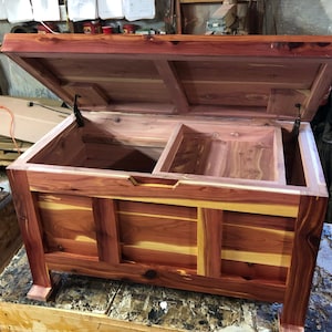 May include: A cedar wood chest with a hinged lid and a divided interior. The chest is made of natural wood with a reddish-brown colour and a light wood grain pattern. The chest is open and the lid is propped up.