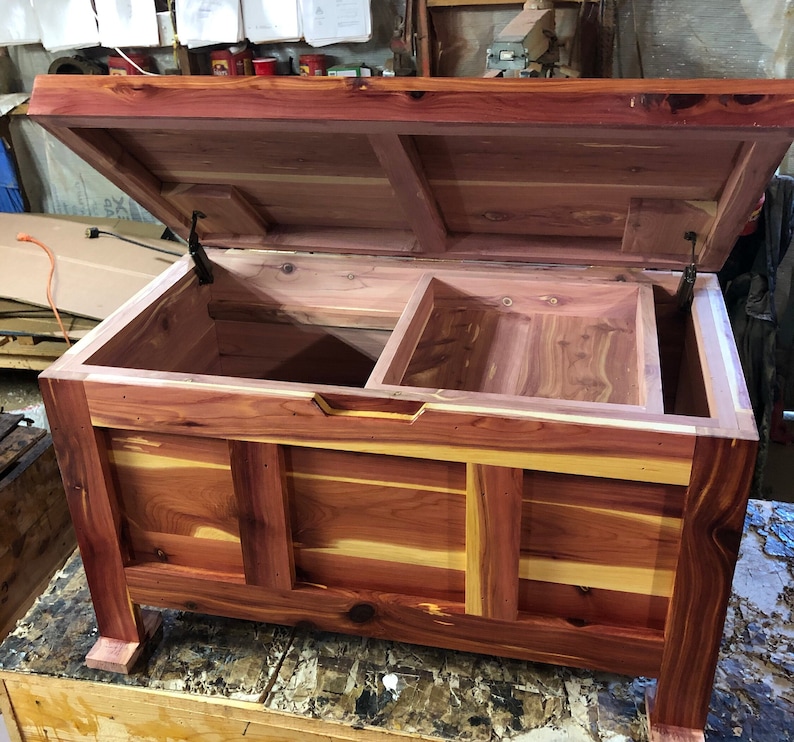 May include: A cedar wood chest with a hinged lid and a divided interior. The chest is made of natural wood with a reddish-brown colour and a light wood grain pattern. The chest is open and the lid is propped up.