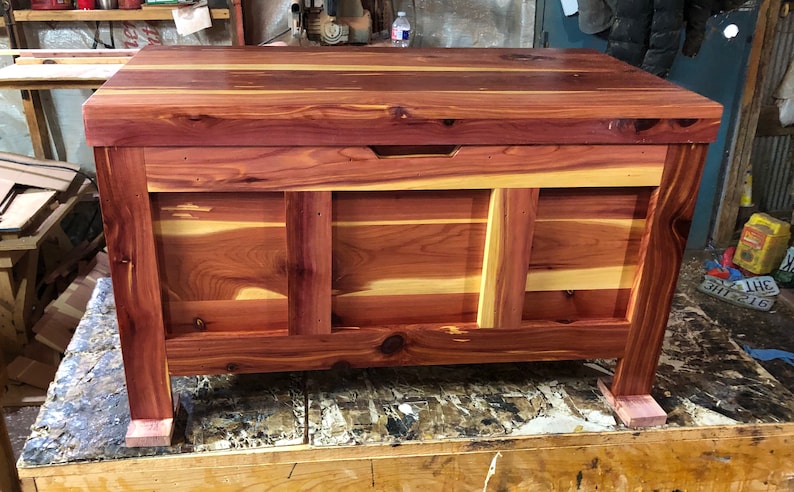 May include: A large cedar chest made from natural wood with a reddish-brown colour and a light wood grain pattern. The chest has a hinged lid and a simple design. It is rectangular in shape with a flat top and a slightly rounded bottom. The chest is sitting on a wooden surface.