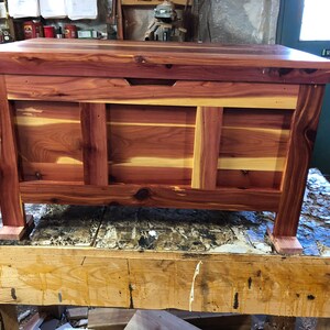 May include: A cedar wood chest with a hinged lid and a small handle. The chest is made of natural cedar wood with a reddish-brown colour and a light brown grain pattern. The chest is sitting on a wooden table.