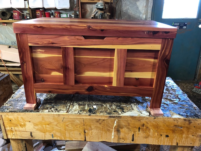 May include: A cedar wood chest with a hinged lid and a small handle. The chest is made of natural cedar wood with a reddish-brown colour and a light brown grain pattern. The chest is sitting on a wooden table.