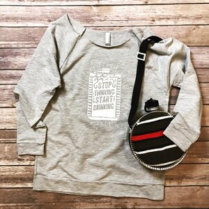 May include: A light gray sweatshirt with the text "STOP THINKING START DRINKING" in white. A black and white striped canteen with a red stripe and a black strap is next to the shirt.