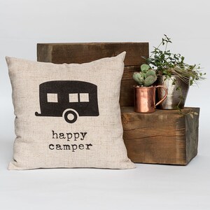 May include: A beige square pillow with a black camper graphic and the words "happy camper" printed below. The pillow is displayed on a wooden block with copper and silver metal planters and greenery.