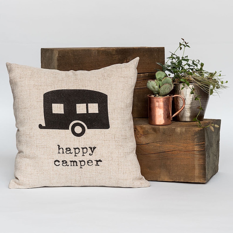 May include: A beige square pillow with a black camper graphic and the words "happy camper" printed below. The pillow is displayed on a wooden block with copper and silver metal planters and greenery.