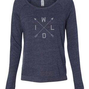 Puede incluir: Una sudadera azul marino de manga larga con la palabra "WILD" en un dise&ntilde;o de flechas cruzadas. El texto y las flechas son de color gris claro. La sudadera tiene cuello redondo y mangas ragl&aacute;n.