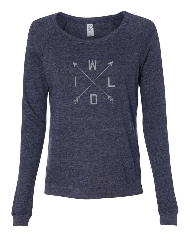 Puede incluir: Una sudadera azul marino de manga larga con la palabra "WILD" en un dise&ntilde;o de flechas cruzadas. El texto y las flechas son de color gris claro. La sudadera tiene cuello redondo y mangas ragl&aacute;n.