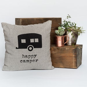 May include: A square, light gray pillow with a black camper graphic and the words "happy camper" printed below. The pillow is displayed on a wooden block with copper and silver metal planters and greenery.