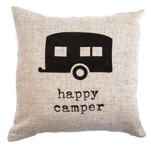 May include: A square beige pillow with a black camper graphic and the words "happy camper" printed below. The pillow has a textured appearance, suggesting a linen or similar fabric. The camper graphic features windows and a wheel.