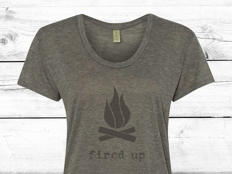 Puede incluir: Camiseta gris jaspeado con cuello redondo y un gr&aacute;fico gris oscuro de una fogata con llamas y las palabras "fired up" impresas debajo. La camiseta est&aacute; hecha de un material suave.