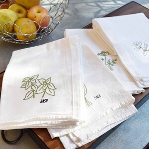 May include: White kitchen towels with green herb illustrations and labels. The towels feature designs of basil, sage, and oregano. A metal basket of fruit sits in the background.