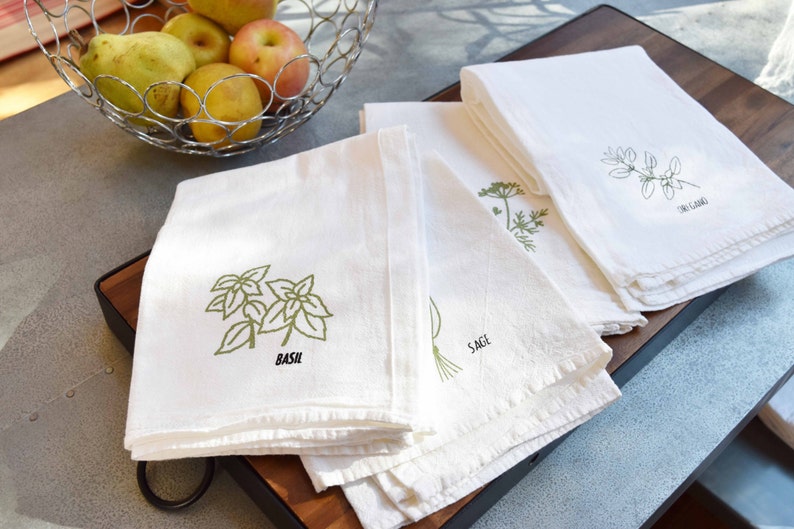 May include: White kitchen towels with green herb illustrations and labels. The towels feature designs of basil, sage, and oregano. A metal basket of fruit sits in the background.