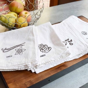May include: Four white kitchen towels with black illustrations of vanilla, ginger, garlic, and an unknown ingredient. The towels are stacked on a wooden cutting board. A metal wire basket with apples and pears sits in the background.