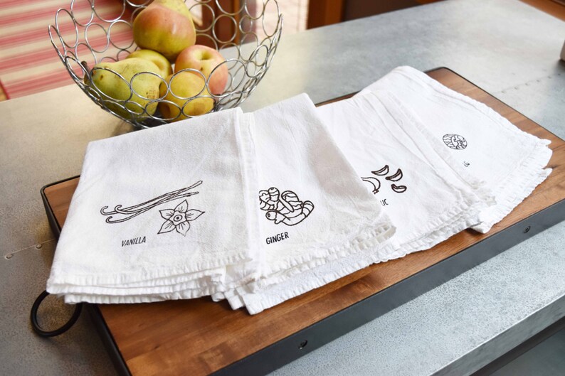 May include: Four white kitchen towels with black illustrations of vanilla, ginger, garlic, and an unknown ingredient. The towels are stacked on a wooden cutting board. A metal wire basket with apples and pears sits in the background.