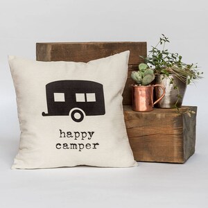 May include: Cream-colored square pillow with a black camper graphic and the words "happy camper" printed below. The pillow is placed on a wooden block with plants and a copper mug in the background. A decorative item for a home.