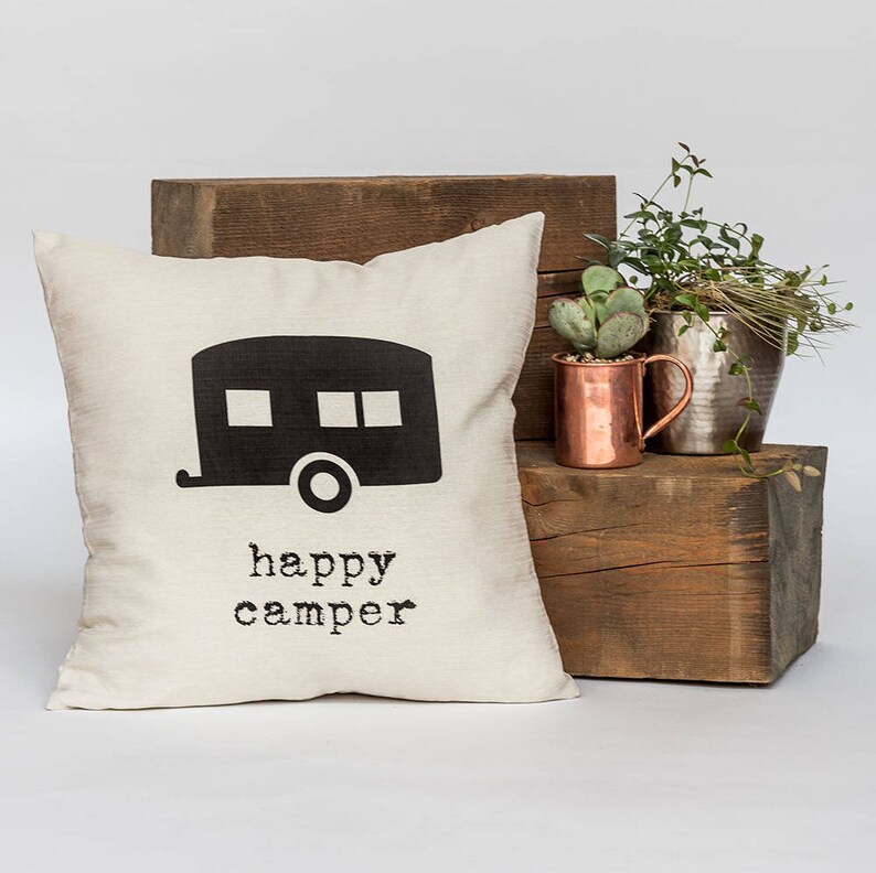 May include: Cream-colored square pillow with a black camper graphic and the words "happy camper" printed below. The pillow is placed on a wooden block with plants and a copper mug in the background. A decorative item for a home.