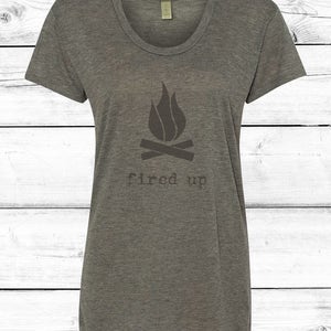 Puede incluir: Una camiseta gris jaspeado con cuello redondo con un gr&aacute;fico de fogata. El gr&aacute;fico incluye un dise&ntilde;o de llama sobre dos troncos cruzados, con las palabras "fired up" impresas debajo en una fuente sencilla. La camiseta es de material suave.