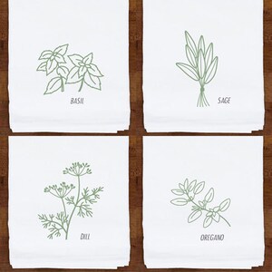 May include: Four white kitchen towels with green line drawings of herbs: basil, sage, dill, and oregano. Each towel has the herb's name printed below the illustration. The towels are displayed on a dark wood surface.