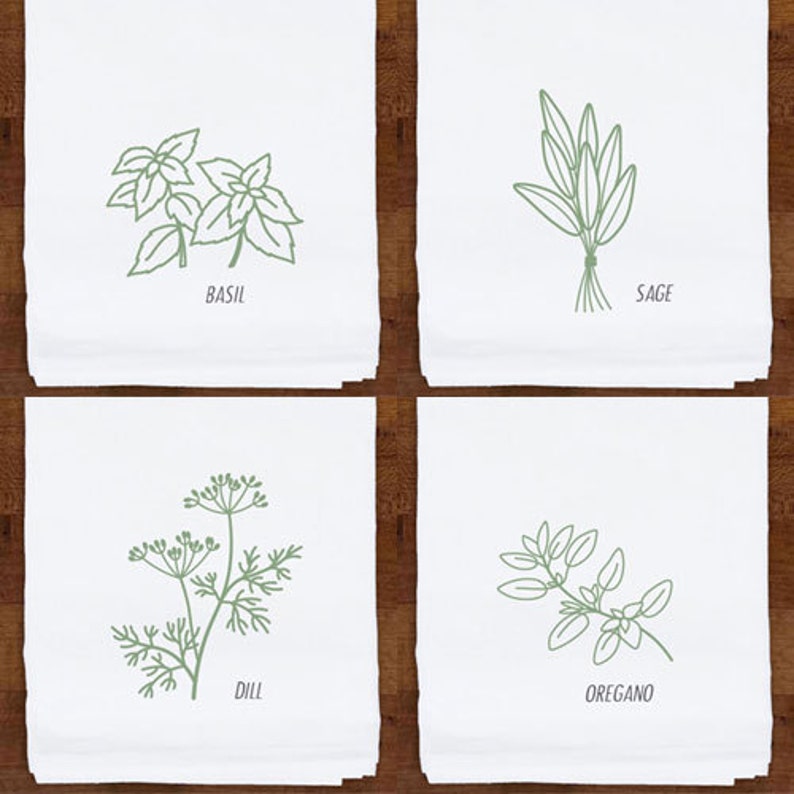 May include: Four white kitchen towels with green line drawings of herbs: basil, sage, dill, and oregano. Each towel has the herb's name printed below the illustration. The towels are displayed on a dark wood surface.