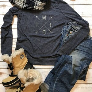 Puede incluir: Un atuendo informal con una camiseta de manga larga azul marino con la palabra "WILD" en un dise&ntilde;o de cruz, vaqueros azules, una bufanda a cuadros en blanco y negro y botas de invierno color canela.