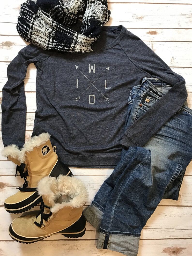 Puede incluir: Un atuendo informal con una camiseta de manga larga azul marino con la palabra "WILD" en un dise&ntilde;o de cruz, vaqueros azules, una bufanda a cuadros en blanco y negro y botas de invierno color canela.