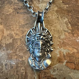 Op de afbeelding: Een zilveren hangerketting met een gedetailleerd maskerontwerp. De hanger toont een gezicht met een kralenmasker en een halo-achtige kroon. De ketting is ook zilver. De hanger is ongeveer 5 cm lang.