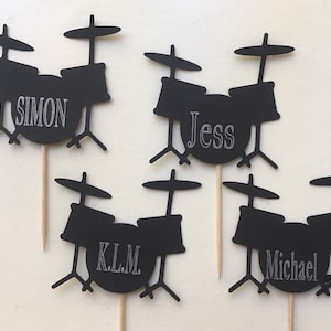 May include: Four black silhouette drum set cake toppers with the names Simon, Jess, K.L.M., and Michael written on the bass drum.
