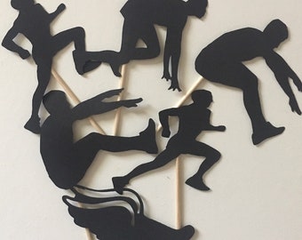 May include: Five black paper silhouettes of athletes in different running poses, each attached to a wooden stick. The silhouettes are arranged in a circle on a white background.