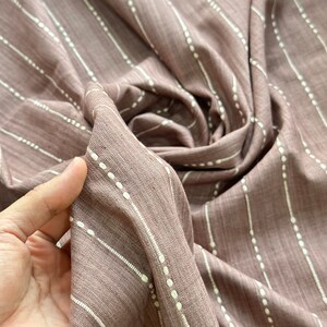 May include: A close-up of a mauve fabric with white stripes and small white dots. The fabric is soft and has a subtle texture.