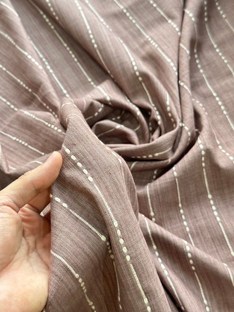 May include: A close-up of a mauve fabric with white stripes and small white dots. The fabric is soft and has a subtle texture.