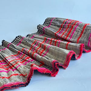 Op de afbeelding: Een geplooide stoffen bies met een witte achtergrond en roze, rode en zwarte strepen. De bies heeft een rafelige rand.