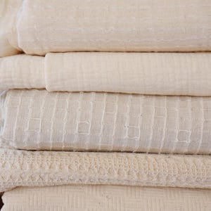 May include: A stack of folded, off-white blankets with varying textures. The blankets are made of natural materials and feature designs like woven squares, horizontal lines, and a lace-like pattern. The stack rests on a woven, brown surface.